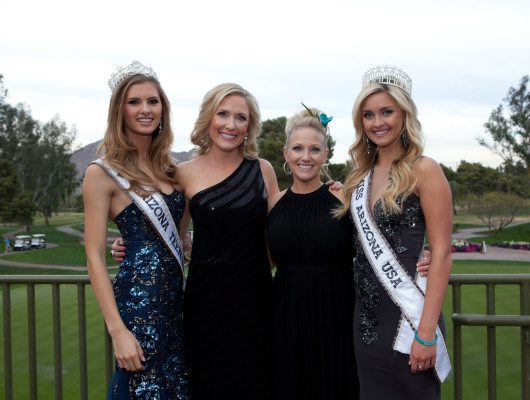 Four women smiling in formal attire outdoors.